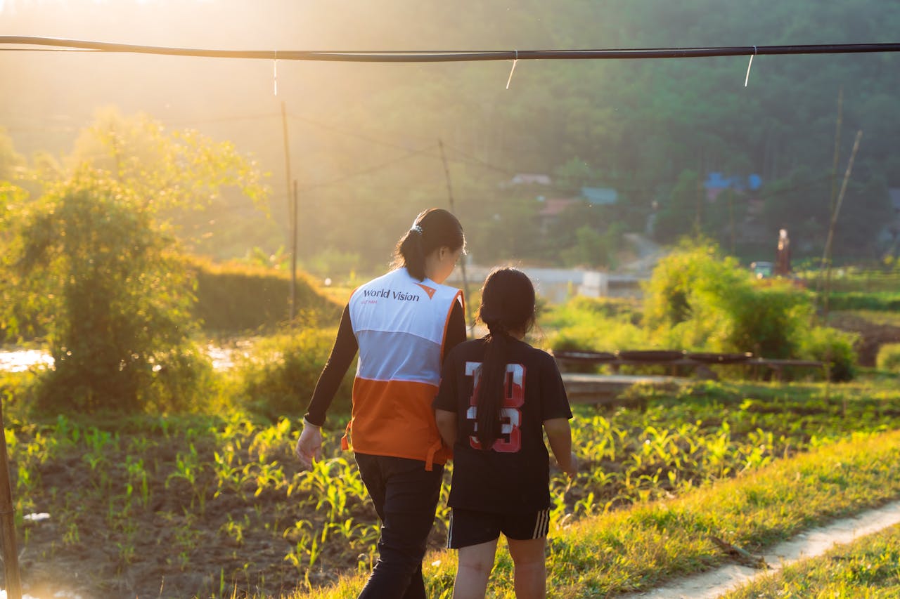 Volunteer in World Vision vest walking with child in scenic rural landscape at sunset.